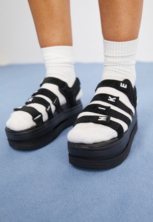 Stopy w białych skarpetkach i czarnych sandałach na platformie z regulowanymi paskami, na których widnieje napis "NIKE", stojące na jasno niebieskim dywanie.