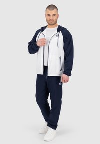 Giacca atletica blu navy e bianca con cappuccio, zip frontale e polsini elasticizzati, abbinata a pantaloni jogger blu navy e sneakers bianche.