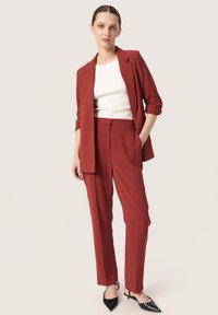 Costum roșu croit, cu o jachetă cu un singur rând de nasturi și pantaloni drepți. Include un top alb cu dungi și pantofi cu vârf ascuțit negri.
