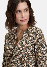 Bluse mit Blumenmuster in Beige mit schwarzen Designs. Weicher Stoff, V-Ausschnitt, geraffte Details an den Ärmeln und aufwendige geometrische Muster.