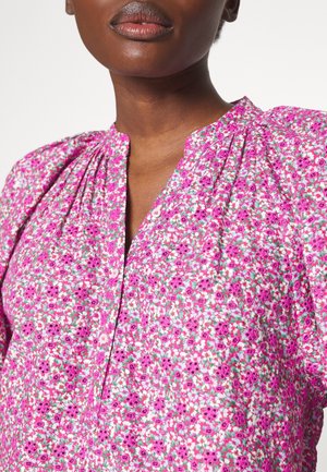 Vrouw draagt een roze blouse met bloemenprint, knoopsluiting, pofmouwen en een geplooide halslijn.