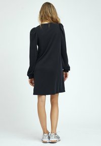 Svart långärmad klänning med en avslappnad passform, som har uppblåsta axlar och knälång längd, i kombination med silverfärgade sportskor.