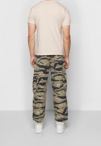 Muž v béžovém tričku, maskáčových kapsáčích s mnoha kapsami a bílých teniskách, stojí čelem k jednoduché šedé zdi.