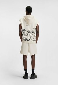 Beige mouwloze hoodie met een zwart boksgrafiek op de achterkant, gecombineerd met beige shorts. Zwarte sokken en schoenen maken de outfit compleet.