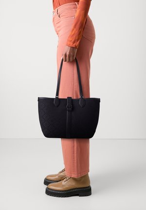 Personne portant un pantalon corail et un haut à manches longues orange, tenant un sac fourre-tout noir à motifs avec double poignées, et portant des chaussures compensées beige.