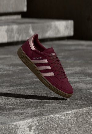 Sneaker Adidas Spezial bordeaux con strisce rosa e suola in gomma che levita sopra gradini in cemento in ambiente esterno.