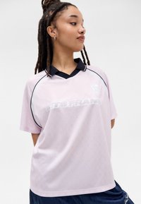 Ροζ κοντομάνικο πόλο μπλουζάκι με μαύρο γιακά και πλευρικές λεπτομέρειες, με υφή και λευκό κείμενο "ETS FRANS" μπροστά.