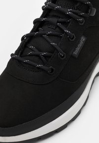 Gros plan sur une chaussure Timberland noire avec une languette en tissu texturé, des lacets noirs à pois blancs et une semelle en caoutchouc blanche.