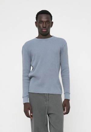 Maglietta a maniche lunghe in maglia waffle azzurro chiaro con colletto tondo e polsini a costine, indossata con pantaloni grigi a vestibilità comoda. Sfondo neutro.