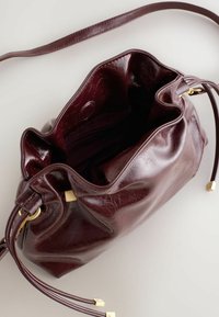 Burgundy nahka nyörilaukku, jossa on teksturoitu pinta. Varustettu kultasävyisillä metalliosilla ja tilavalla sisäosastolla, jossa on pieni vetoketjukuitti.