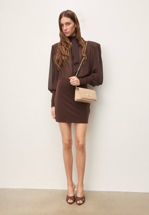 Brązowa bluzka z długim rękawem i wysokim kołnierzem, zestawiona z dopasowaną brązową mini spódnicą. Dodatki to mała beżowa torebka i brązowe sandały na obcasie odkryte na palcach.