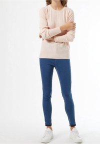 Lichtroze gebreide trui met een ribbelstructuur, gecombineerd met blauwe skinny jeans en witte sneakers, staand tegen een effen achtergrond.