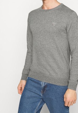 Uomo che indossa un maglione grigio aderente a maniche lunghe con un piccolo logo sul petto, abbinato a jeans in denim blu, in piedi contro uno sfondo bianco.
