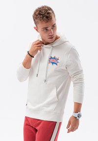 Sudadera blanca con un gráfico colorido de "¡OMG!" en el pecho, con cordones y un bolsillo delantero, combinada con pantalones rojos y un reloj.