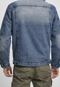 Hombre vistiendo una chaqueta de mezclilla azul desgastada y pantalones cargo verdes oliva, mostrado desde la espalda contra un fondo claro y liso.