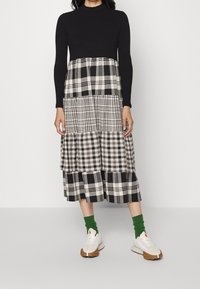 Top negro de manga larga con una falda de capas que presenta diferentes patrones de cuadros en blanco y negro. Acompañado de zapatillas blancas y calcetas verdes.