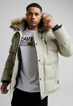 Jeune homme ajustant la capuche d'une doudoune beige clair avec bordure en fourrure, portant un t-shirt blanc imprimé et un pantalon noir.