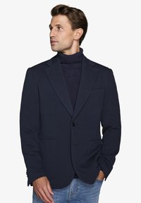Marineblazer lavet af blødt stof, med en spids revers, én-knap lukning og to frontlommer, lagdelt over en marineblå turtleneck.