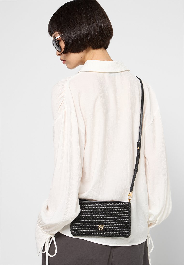 Kobieta z krótkimi ciemnymi włosami nosząca duże okulary przeciwsłoneczne, białą bluzkę i trzymająca małą czarną torebkę typu crossbody o fakturze.