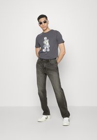 Tricou gri cu un grafic de urs, asortat cu blugi gri închis. Modelul poartă adidași albi cu accente verzi și ochelari de soare.