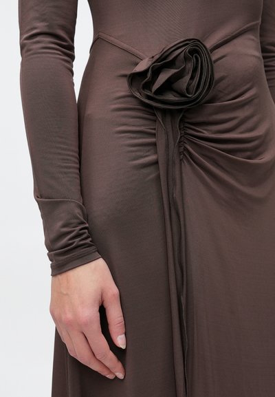 Κλείσιμο εικόνας ενός χεριού που ακουμπάει στο πλάι ενός μακρυμάνικου καφέ φορέματος με ραμμένο ύφασμα και διακοσμητικό ροζέτα στη μέση.