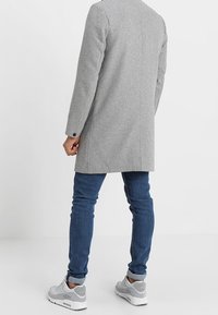 Cappotto di lana grigio con un design elegante, caratterizzato da una singola fessura posteriore, abbinato a jeans blu e sneaker grigie. Texture liscia e vestibilità su misura.