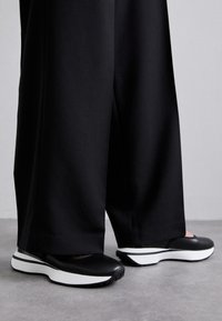 Schwarze Slipper mit glatter Oberfläche und weißer Sohle. Mit runder Zehenform und subtilen Akzenten nahe der Ferse.