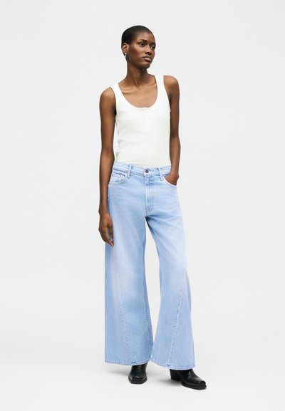 Lihavõitu naine, kes kannab valget varrukateta toppi, helesiniseid laiapõlvalisi teksaseid ja musti pahkluu saapaid, seistes ühe käega pihikusse pistetuna.