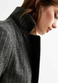 Chaqueta de mezcla de lana gris con un cuello alto y textura de rayas verticales, que presenta una silueta estructurada y un acabado suave.