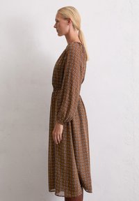 Bruine jurk met patroon, lange mouwen, een elastische taille en een middellange snit. Heeft een geometrisch ontwerp in lichtere tinten.
