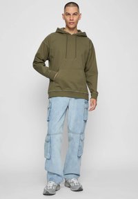 Olívazöld kapucnis pulóver elejében zsebbel, világoskék cargo nadrággal, amely több zsebbel és laza szabással rendelkezik.