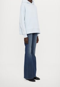 Svetle modrá oversized mikina s kapucí a přední kapsou, s bílým logem na kapuci, spárovaná s rozšířenými tmavými džíny a černými botami.