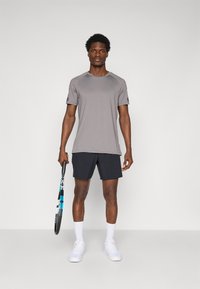 T-shirt de sport gris à manches courtes, associé à un short noir. Un homme debout tient une raquette de tennis, portant des chaussettes et des chaussures blanches.