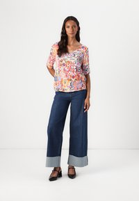 T-shirt colorato con motivo, scollatura a V e maniche corte, abbinato a pantaloni in denim scuri a gamba larga con risvolti a contrasto e scarpe nere.
