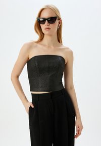 Sort strapless crop top med en tekstureret overflade; parret med højtaljede sorte bukser. Model iført sorte katteøje-solbriller.