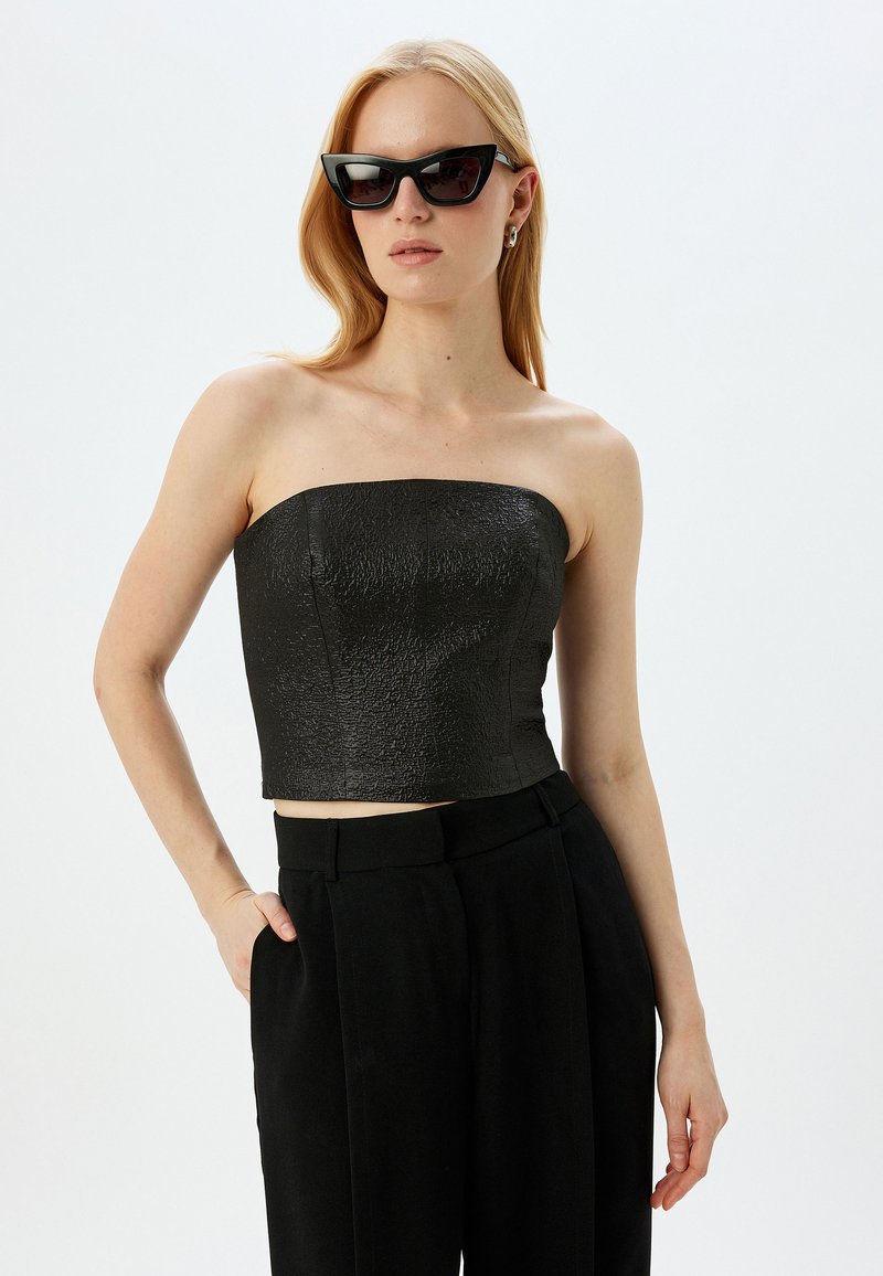 Sort strapless crop top med en tekstureret overflade; parret med højtaljede sorte bukser. Model iført sorte katteøje-solbriller.