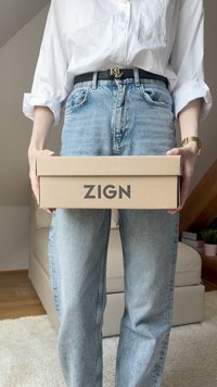 Scatola di cartone marrone con "ZIGN" stampato in nero sul davanti, tenuta in mano, con jeans azzurri chiari usurati e camicia bianca a bottoni visibili.