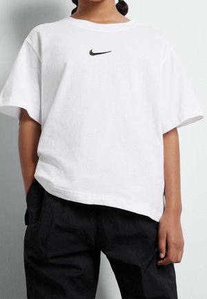 Person iført en løs hvid Nike t-shirt med en sort swoosh-logo centreret på brystet og sorte bukser, stående mod en lys baggrund.