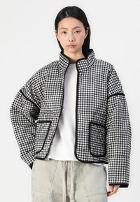 Jachetă puffer quilted, cu un model în carouri alb-negru, având un guler înalt, două buzunare frontale și un tiv contrastant negru.