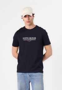 Marineblå bomulds T-shirt med "TOMMY HILFIGER" logo i hvid og rød. Sæt sammen med lyseblå denim jeans og en beige kasket.