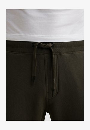 Ceinture de pantalon de survêtement vert foncé avec cordon de serrage noué en un nœud, associée à une chemise blanche légèrement rentrée.