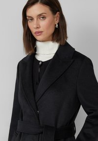 Cappotto nero in misto lana con colletto a punta, vita con cintura e chiusura a bottoni, indossato sopra un maglione bianco a coste con collo alto e un maglione nero.
