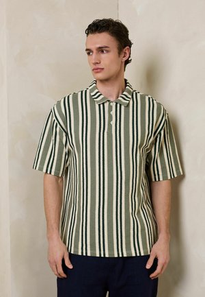 Fiatal férfi, aki rövid ujjú, galléros inget visel függőleges zöld és fehér csíkokkal, egy bézs fal mellett áll, balra néz.