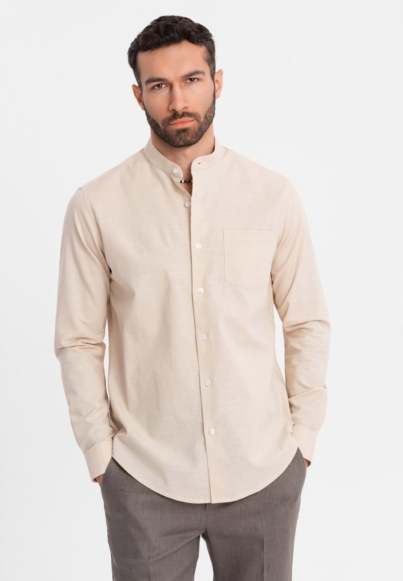 Homme aux cheveux foncés et barbe portant une chemise beige boutonnée à col mandarin et un pantalon gris, debout les mains dans les poches.