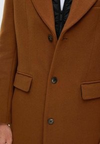 Brun ullblanding frakk med spiss krage, to frontlommer og tre svarte knapper; innsiden har en kviltet svart foring.