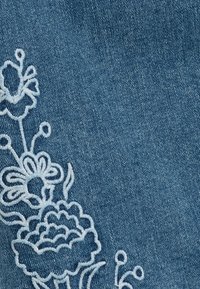 Tejido de mezclilla en azul con bordado floral blanco a lo largo de un lado, con intrincados diseños de flores y hojas. Textura suave.