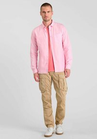 Camicia a righe rosa, con bottoni, indossata sopra una t-shirt color corallo, abbinata a pantaloni cargo beige e sneakers bianche. Outfit casual con tasche funzionali.