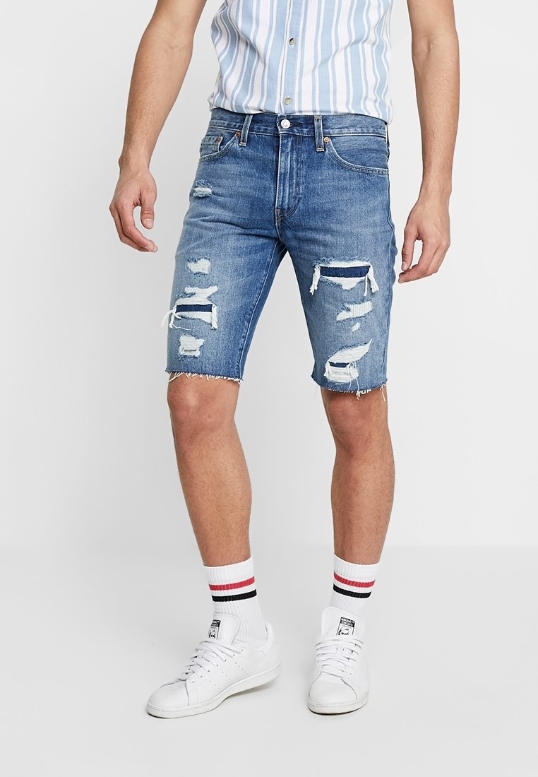 Homme portant un short en jean bleu déchiré, une chemise rayée à boutons, des baskets blanches et des chaussettes blanches avec des rayures rouges et noires, debout sur un sol blanc.