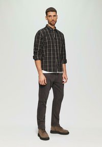 Chemise à carreaux noire et beige pour homme, portée sur un t-shirt blanc, associée à un jean gris foncé et des bottes lacées marron à la cheville.