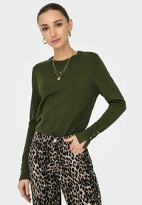 Grüner Strickpullover mit langen Ärmeln und Knopfdetails an den Bündchen, kombiniert mit beigefarbenen und schwarzen Leopard-Muster-Walkhosen.